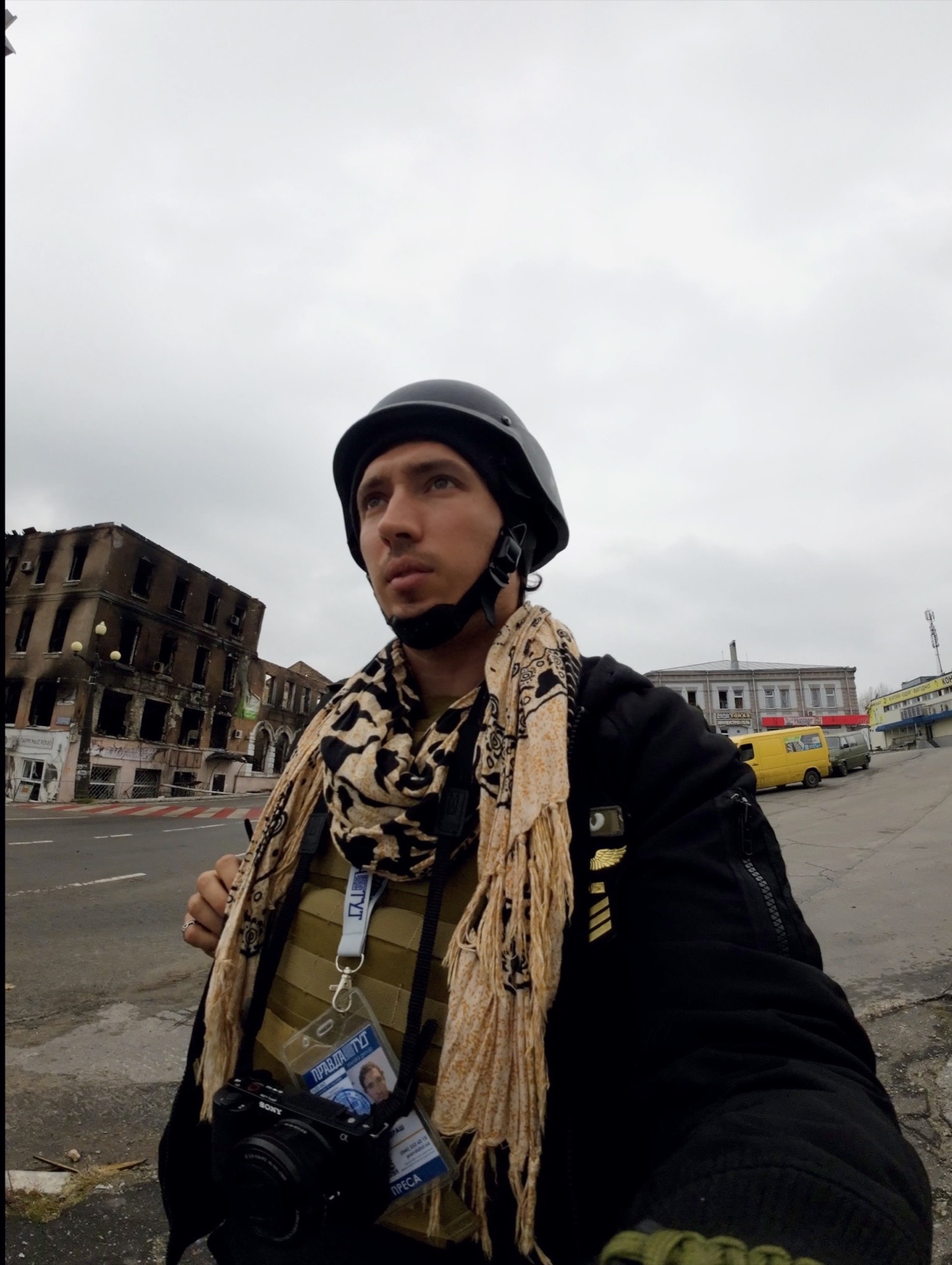 Emanuel in a helmet and body armor with a press credential, standing in front of a fire-gutted building in Kupiansk